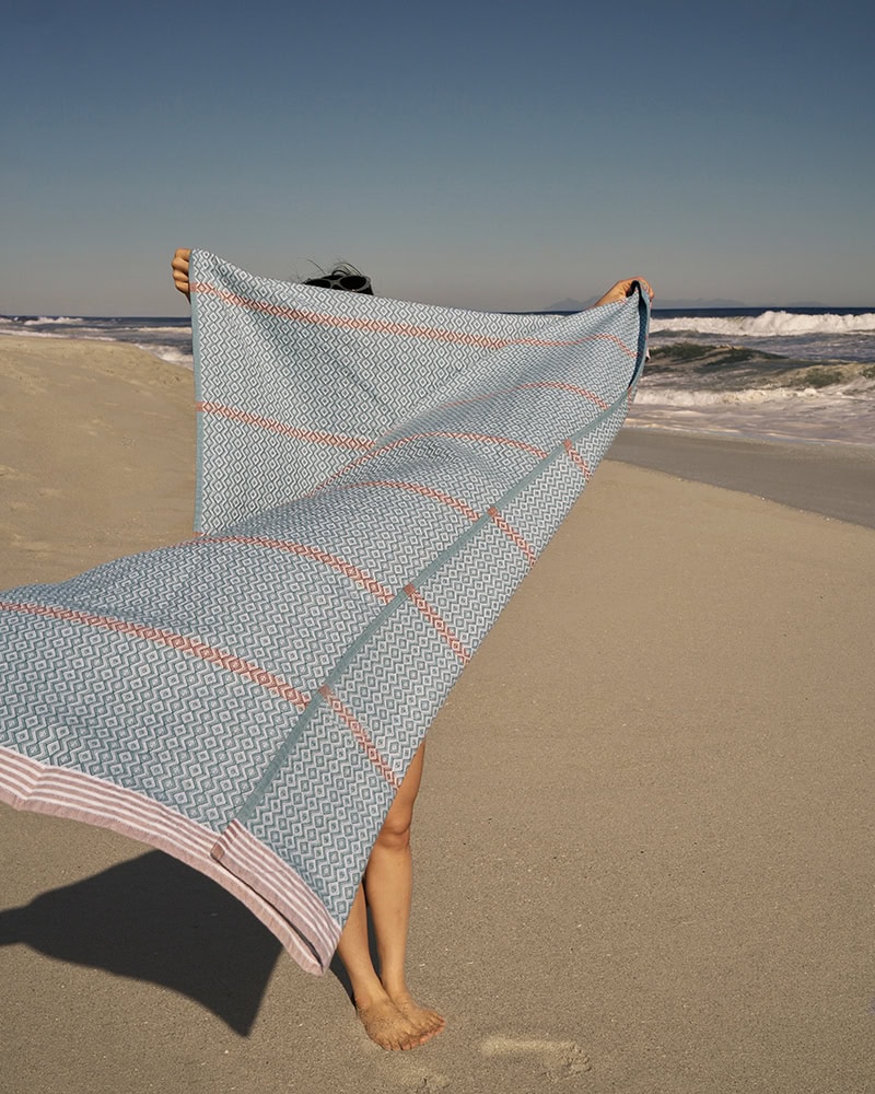 Model on the beach flicking the Itawuli Bath Towel in Dusty Teal